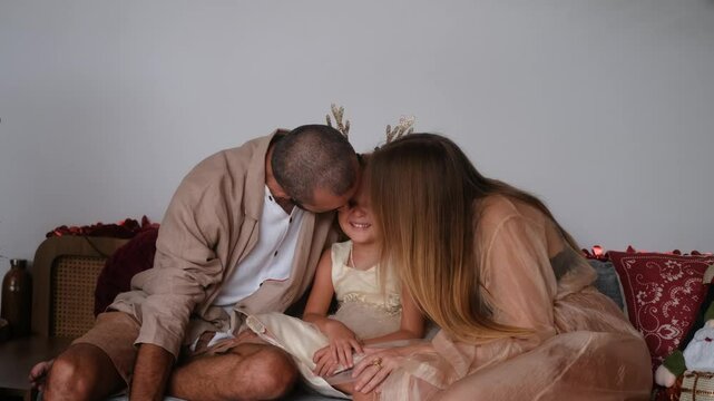 Family Christmas. A little girl sits in the middle between her parents with a toy poodle in her arms. Mom and Dad kiss their daughter from both sides at the same time, she laughs and happy.