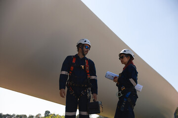 A couple of electrical engineers examine the blades of a wind turbine, standing at the base of a...