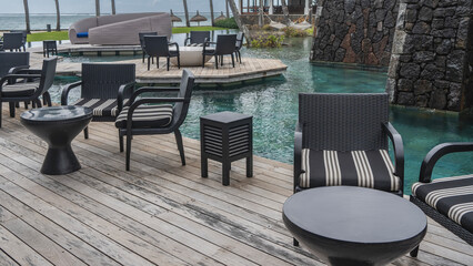 The recreation area at the resort by the swimming pool. There are wicker chairs with soft cushions, round tables on the board platform. Clear turquoise water. In the distance - the ocean, lawn, palms