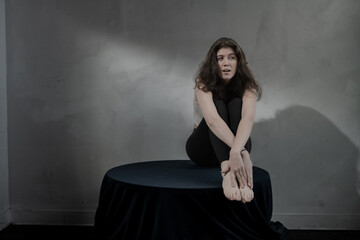 The woman sits on a draped round table, her feet and hands closely positioned in front of her, with...