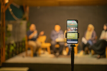 A smartphone mounted on a tripod live streaming a panel discussion in a cozy indoor setting, highlighting modern technology, connectivity, and innovative event broadcasting techniques.