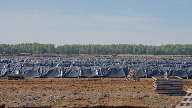 Panoramic view of peat field under bright sky, vast harvesting area with raw materials covered up by black tarps stretching to edge of forest
