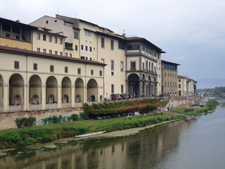 Fototapeta premium The Uffizi Gallery's historic facade overlooking the Arno River, with lush greenery along the riverbank and a serene, cloudy sky in the background