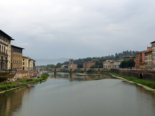  Scenic view of the Arno River surrounded by historic buildings and a bridge under a cloudy sky,...