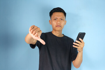 Asian young man posing with a thumbs-down gesture while holding mobile phone 