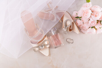 A pair satin beige shoes with a rhinestone bow on beige background with a bouquet of peonies...