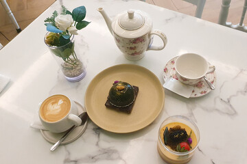 Two delicious cakes, one cup hot cappuccino with latte art and a kettle filled with tea served on the table in a café during lunch break