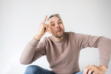 A man sits against a white background, pointing to his head with a playful or insightful...