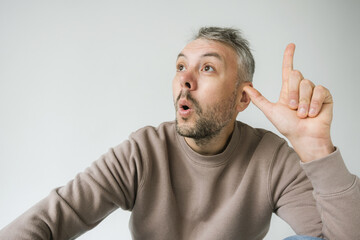 A man sits against a white background, looking up and to the side with a surprised or curious expression. He s holding his hand to his ear, as if listening intently, while his other hand points upward