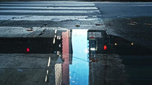 Close-up of puddle in urban downtown environment reflection of buildings and sky as car drives over crosswalk