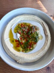 A bowl of hummus with olive oil, small pieces of red pepper and fresh green herbs.  In a white ceramic bowl on a restaurant table. 