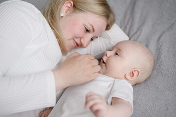 Mom lies on soft bed, next to small child, enjoying tender moment together. Woman smiles, gently strokes baby, lovingly looking at infant. Concept of motherhood, childcare, maternity leave, nanny