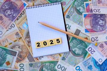 Polish zloty banknotes in the background, a notebook with the inscription ‘2025’ on plastic cubes, and a pencil, a personal finance concept.