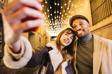 A joyful couple captures a memorable moment with a selfie beneath glowing string lights in an urban setting. 