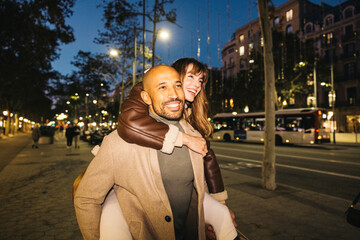 A smiling couple delight in a piggyback ride along a lively city street at night enhancing the romantic evening ambiance.