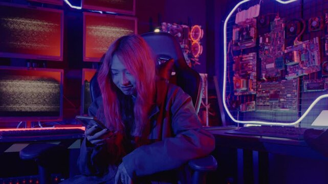 Asian woman in front of a computer monitor display using mobile phone, neon light, Hacker wear black jacket at system control room, Security guard working on multiple monitors together smartphone