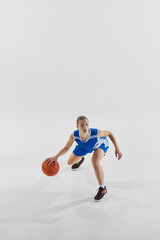 Fototapeta premium Full-length dynamic image of young teen girl, basketball player in blue uniform in motion with ball during game, training against white background. Concept of sport, youth, motion, healthy lifestyle