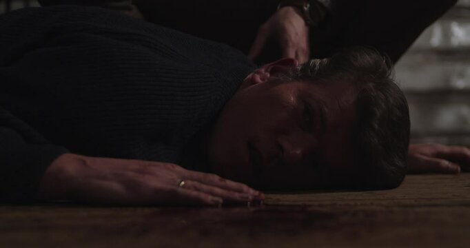 Close up of a dead man lying on a wooden floor with a puddle of blood while a doctor checks his pulse