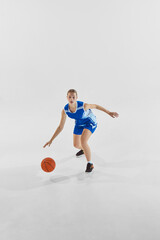 Full-length dynamic image of young teen girl, basketball player in blue uniform in motion with ball during game, training against white background. Concept of sport, youth, motion, healthy lifestyle