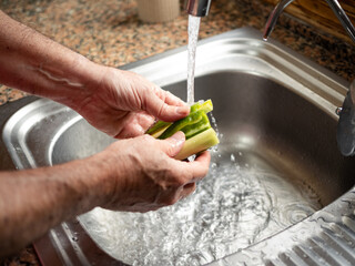 Hands of a man are rinsing fresh green peppers and leek under running water in a kitchen sink. The countertop has a textured surface and the water splashes around, indicating a preparation for cooking