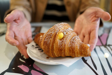 A flaky croissant with custard filling, dusted with powdered sugar, served on a plate. Hands gesture in a welcoming manner, creating a cozy café atmosphere.