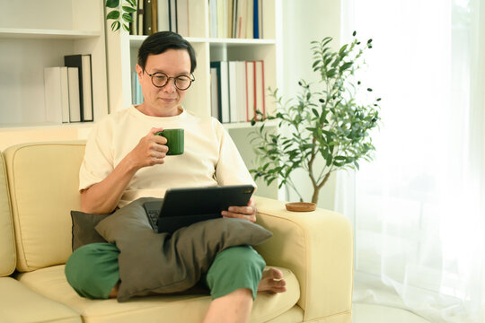 Happy middle aged man sipping coffee from a green mug and browsing on digital tablet - Powered by Adobe