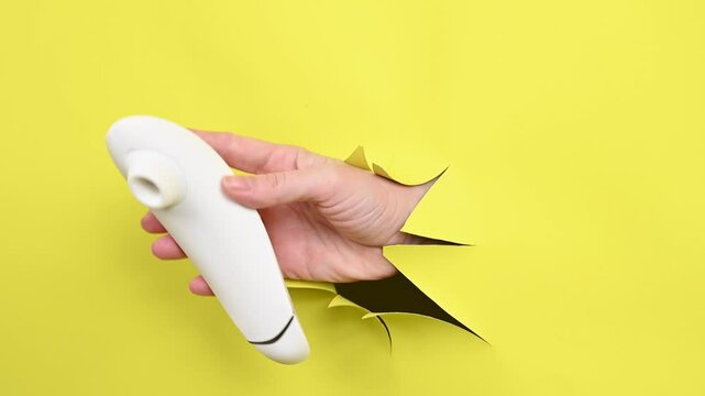 Female hand holding white clitoral vacuum vibrator sticking out of yellow paper background. 