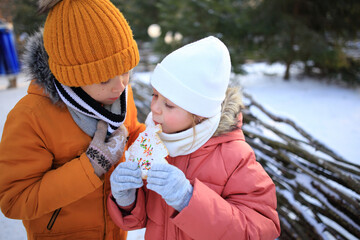 A brother and sister share a delightful moment as they eat gingerbread cookies outdoors against the backdrop of a snowy forest. 