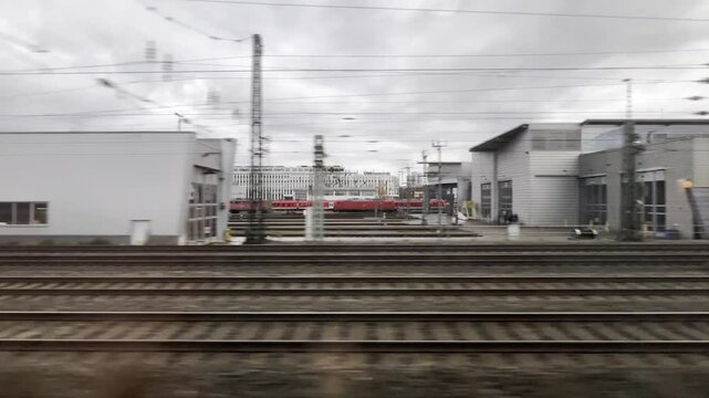View from the window of a train passing a large station. Various buildings and railroad tracks fly by. From the train's window you can see the city and oncoming and passing trains.