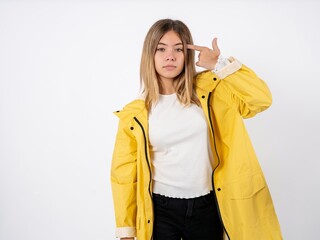 Unhappy Beautiful teenager girl wearing yellow raincoat makes suicide gesture and imitates gun with hand, curves lips keeps two fingers on temple, shoots, being tired of everything,