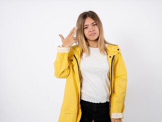 Unhappy Beautiful teenager girl wearing yellow raincoat makes suicide gesture and imitates gun with hand, curves lips, keeps two fingers on temple