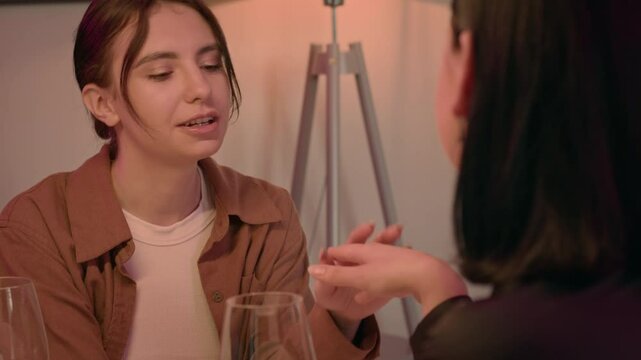 Two women meet in a cafe, focusing on one. One gently touches and strokes her friend's hand, smiles lighting up their faces. They share white wine, clinking glasses in a moment of closeness.