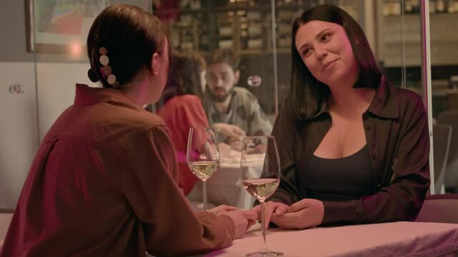 Two women meet in a cafe, sipping white wine and engaging in open flirtation. One brushes her hair behind her ear and places her hand on her partner's, showing clear romantic intent.