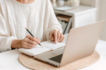 Fototapeta premium Businesswoman writing notes in notebook while working remotely on laptop computer at home office, wearing white blouse, sitting at white table with wicker placemat