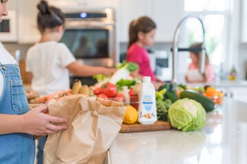 Family unpacking groceries in a bright kitchen, with a focus on a young girl holding a grocery bag while others organize fruits and vegetables.
