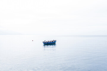 Serene, minimalist scene featuring a single blue boat floating on still water, with birds perched on it. The background is misty, with faint outlines of distant mountains.