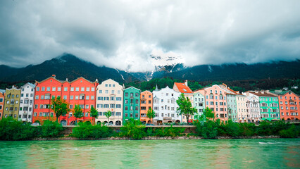 Fototapeta premium Colorful Houses Along a Tranquil River in Innsbruck with Snow-Capped Mountains and Cloudy Skies in the Background