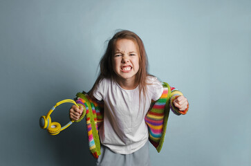 A young girl with long brown hair displays a fierce, angry expression. She s wearing a colorful striped cardigan and holding yellow headphones