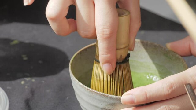 A young Asian woman brews trendy matcha tea following a traditional Japanese recipe. Stirring green tea powder with whisk chasen in a chawan bowl. Tea ceremony, close up.