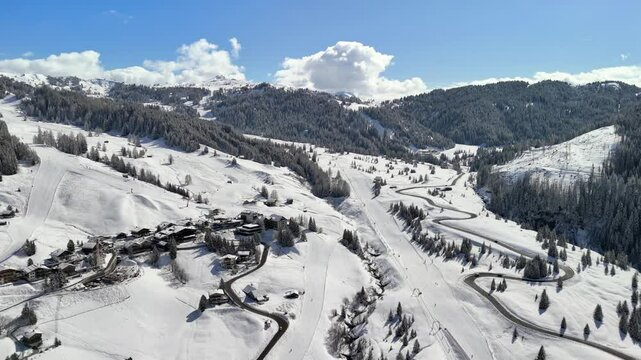 Alta Badia, Italy: Aerial view of ski resort in snowy Dolomites. Charming town landscape with chalets and hotels surrounded by snow-capped peaks, sunny day, blue sky
