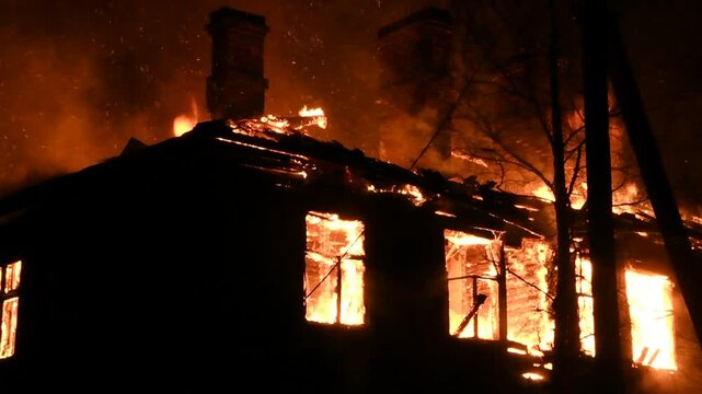 House is on fire at night, flames inside at home, burning windows. The wooden building is ablaze, completely engulfed in fire. Loss of housing, disaster, trouble for unhappy people.