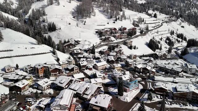 Arabba, Italy: Aerial view of ski resort in snowy Dolomites. Charming town landscape with chalets and hotels surrounded by snow-capped peaks, sunny day, clear blue sky
