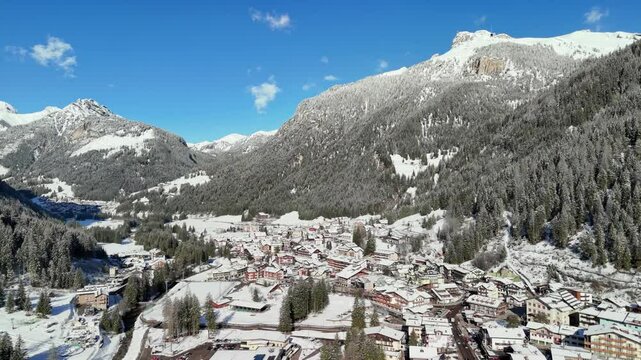 Canazei, Italy: Aerial view of ski resort in snowy Dolomites. Charming town landscape with chalets and hotels surrounded by snow-capped peaks, sunny day, clear blue sky
