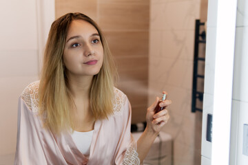 A blonde girl in a pink bathrobe is sprayed with perfume in the bathroom. Beautiful young woman...