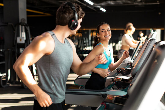 Happy fit man and woman doing cardio workout in modern gym interior, couple running on treadmills and talking with each other