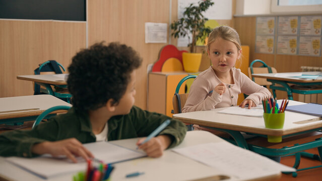 Multiracial diverse little classmates multiethnic students pupils two children kids Caucasian girl help African American boy suggest correct answer to exam writing test talking in school class lesson