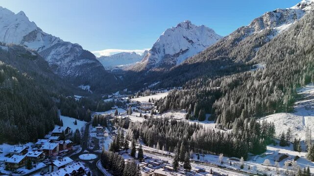 Canazei, Italy: Aerial view of ski resort in snowy Dolomites. Charming town landscape with chalets and hotels surrounded by snow-capped peaks, sunny day, clear blue sky
