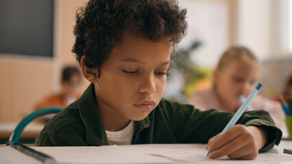 Little African American guy boy writing notes task class lesson primary school write in copybook kid schoolboy son child pupil student learning listen teacher studying education sitting at table desk