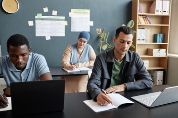 Medium shot of adult multiracial man concentrated on writing task sitting at desk in study group with other students attending language courses class