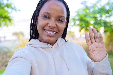 A smiling woman takes a selfie outdoors while waving with her hand. She is dressed in a light...